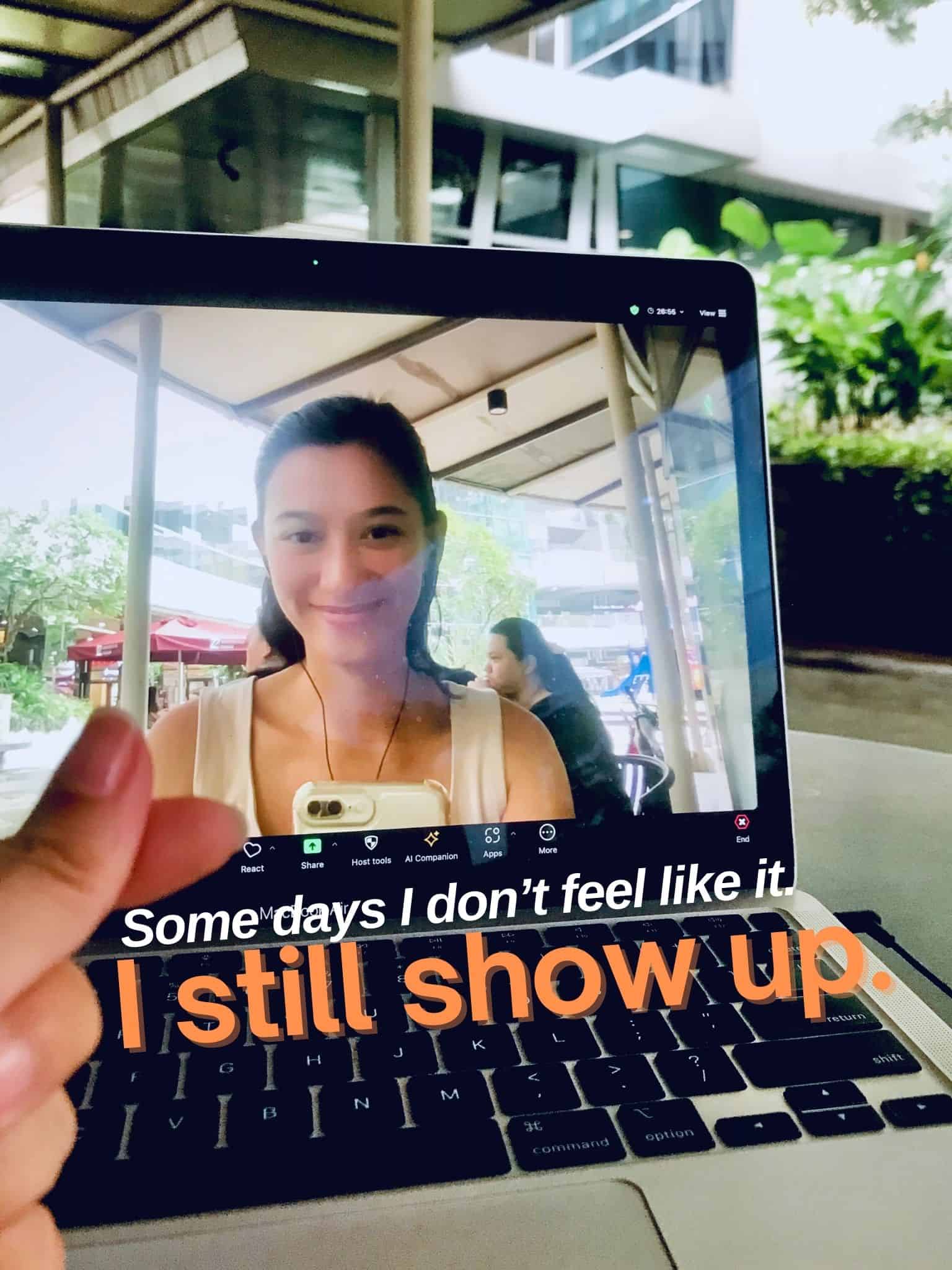 Smiling woman on laptop screen at outdoor café with greenery and modern architecture in background.